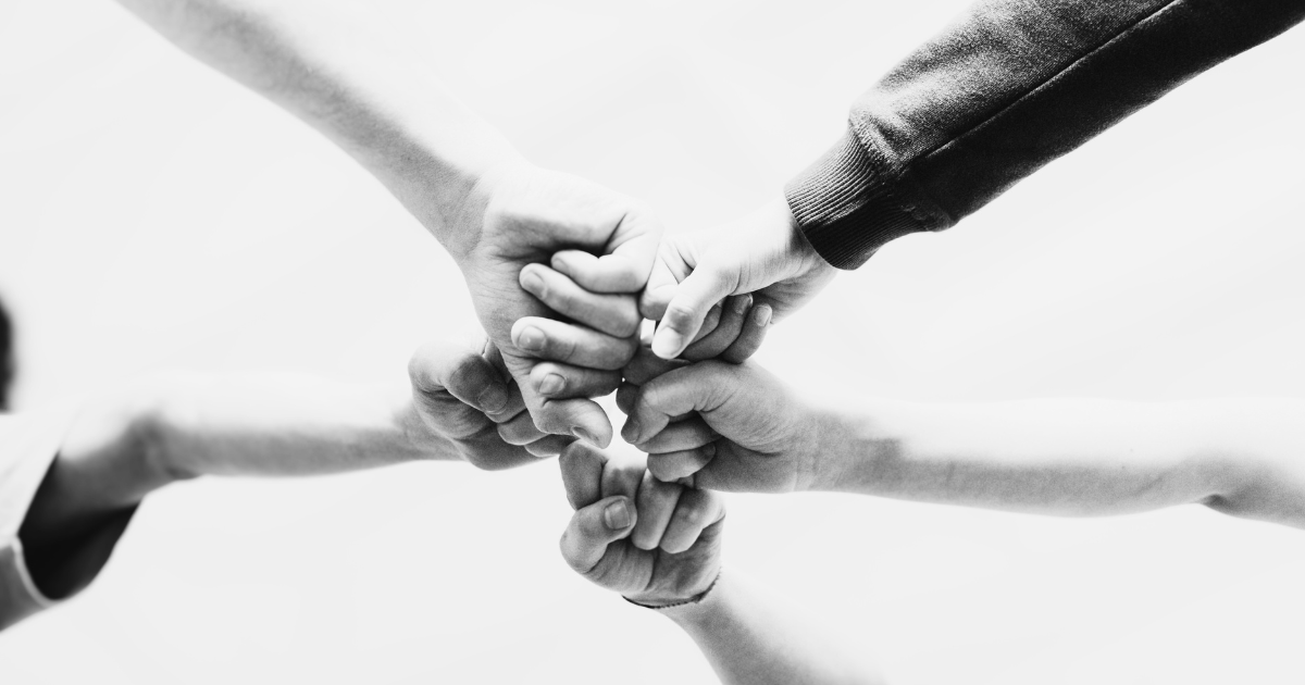 Black and white photo of five peoples forearms all fist bumping to illustrate the effect Qualified Charitable Distributions (QCDs) has on a community.