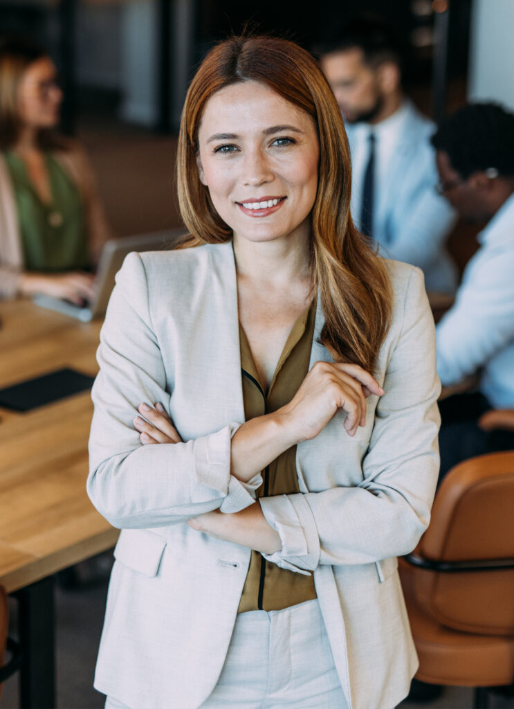 Woman in her 30s standing at a desk with people working behind her to illustrate MedTech professionals we offer our retirement planning services to.